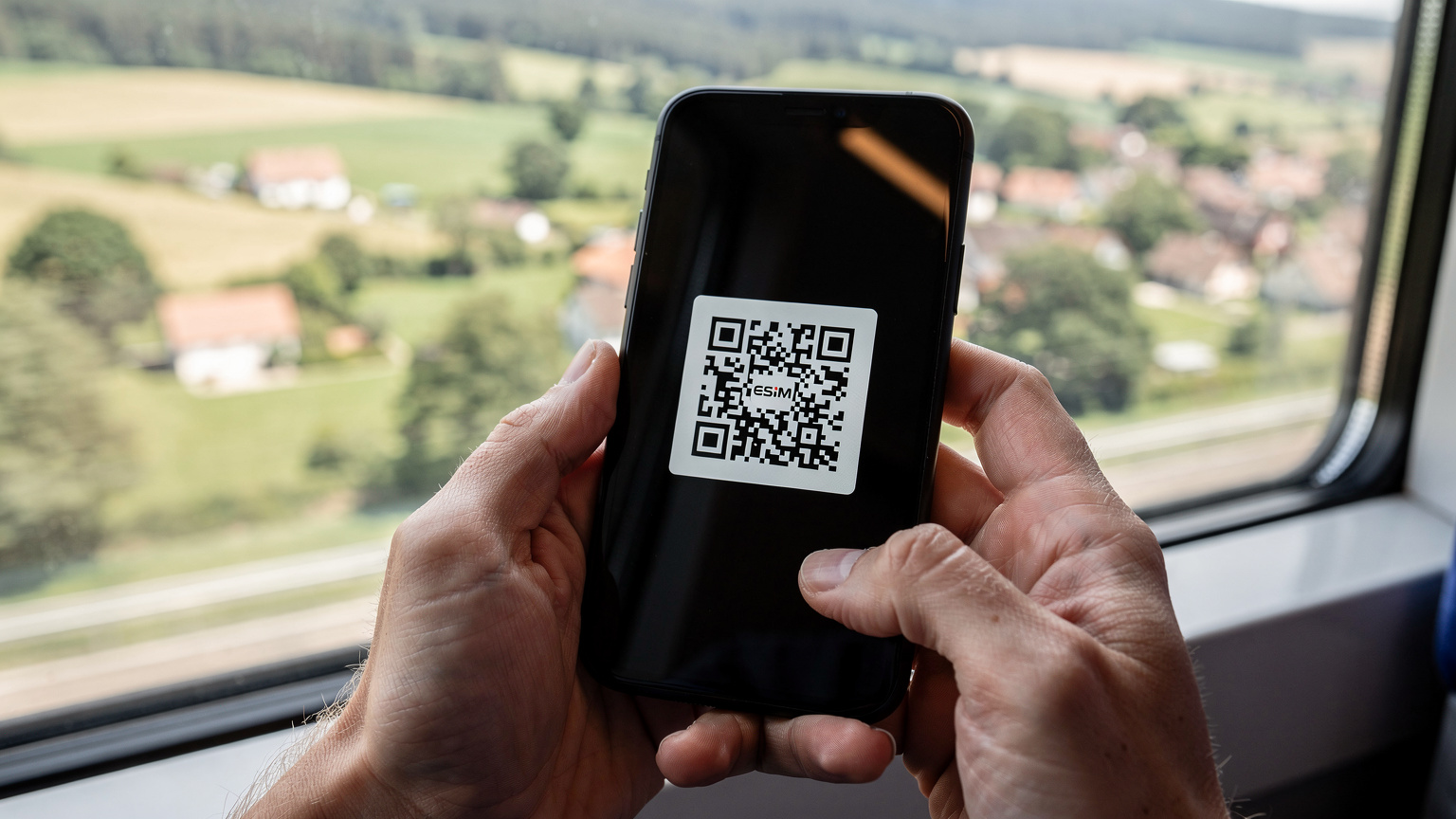Nahaufnahme von Händen, die ein Smartphone halten, auf dessen Bildschirm ein QR-Code für die eSIM-Installation angezeigt wird, mit einer vorbeiziehenden deutschen Landschaft im Hintergrund, gesehen aus einem Zugfenster.