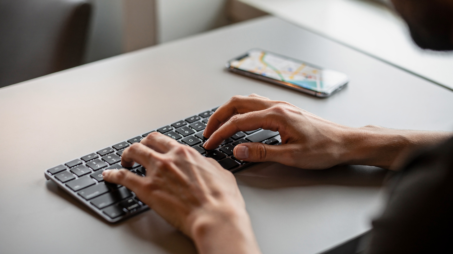 Close-up of a person's hands efficiently typing on a laptop keyboard, with a smartphone displaying a travel map app in the background, suggesting seamless navigation and work.