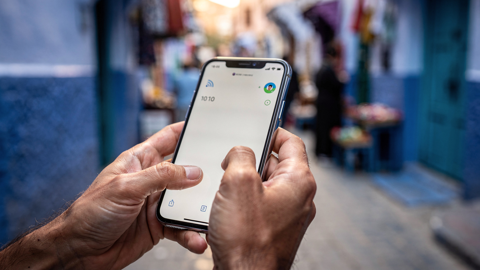 Gros plan sur les mains d'une personne qui manipule son smartphone, l'écran affichant des barres de signal réseau et des icônes de données, sur fond de rue animée à Chefchaouen.