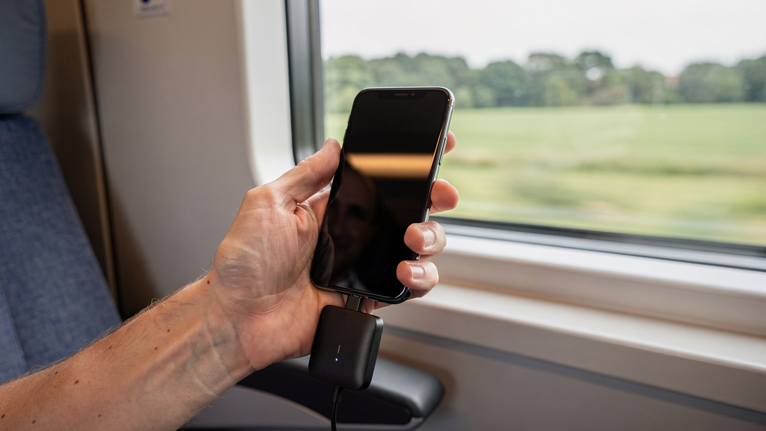Un călător verifică nivelul bateriei la un telefon cu eSIM activ și un power bank conectat, într-un tren de mare viteză în Germania, cu peisajul rural trecând rapid pe fereastră.