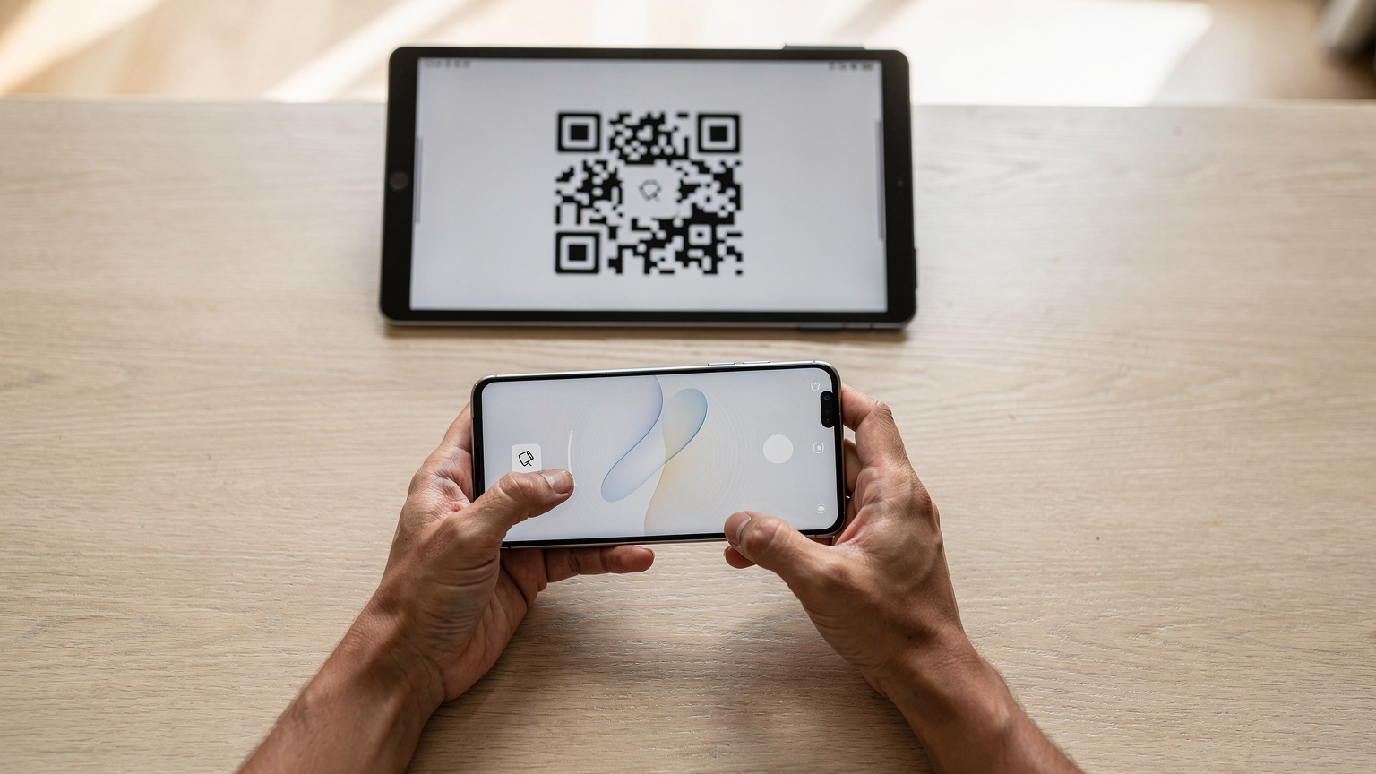 Manos de un adulto latinoamericano sosteniendo un smartphone en primer plano, mostrando la pantalla de configuración de eSIM, mientras escanea un código QR desde una tablet que se ve parcialmente en el fondo. El escenario es el interior de una casa familiar moderna, con una mesa de madera clara y luz natural.
