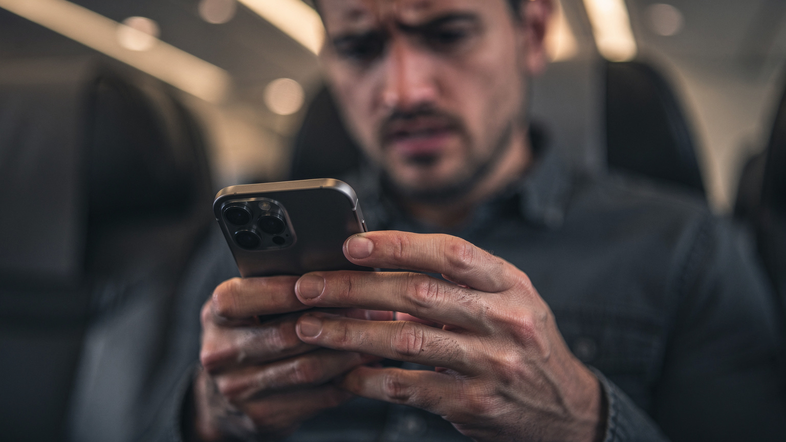 Primer plano de las manos de un viajero con una expresión de frustración, sosteniendo su iPhone 17 y mirando la pantalla con preocupación, en un asiento de avión o en una sala de espera de aeropuerto con poca luz.