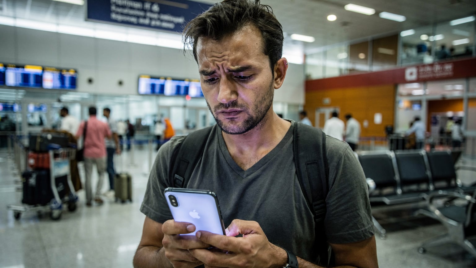 A focused traveler with their phone displaying a 'No Service' message, looking frustrated at an airport arrival gate in Mumbai, India, with other passengers in the background.