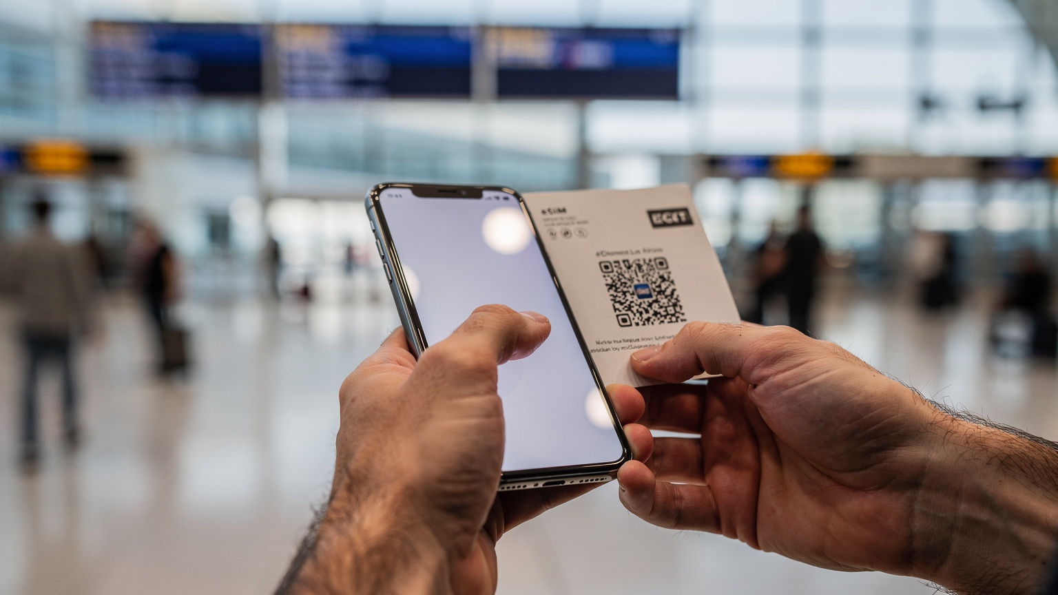 Detailní záběr na ruce cestovatele, který drží chytrý telefon a skenuje QR kód eSIM na letišti v Los Angeles, s rozostřeným pozadím letištního terminálu a cestujících.