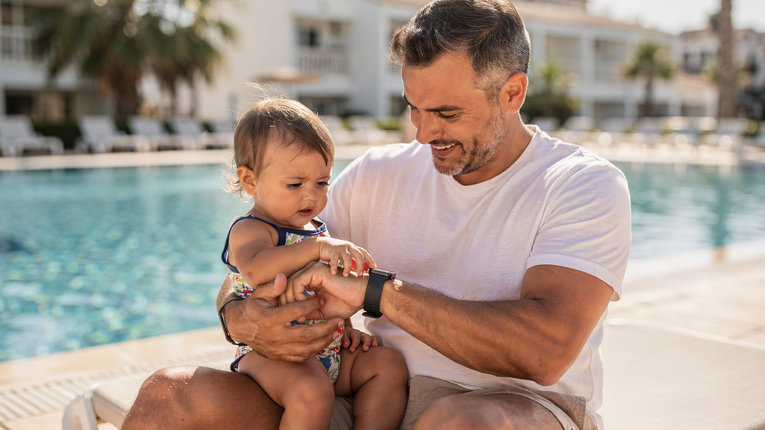 Nahaufnahme eines Vaters, der mit seinem Kleinkind im Arm entspannt am Pool sitzt und auf seine Apple Watch schaut, während das Kind neugierig auf den Bildschirm zeigt.
