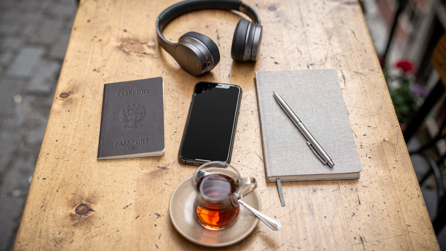 An overhead flat-lay shot of a digital nomad's desk in a café, featuring a smartphone, passport, travel journal, and a cup of coffee, symbolizing preparation and essential travel items.