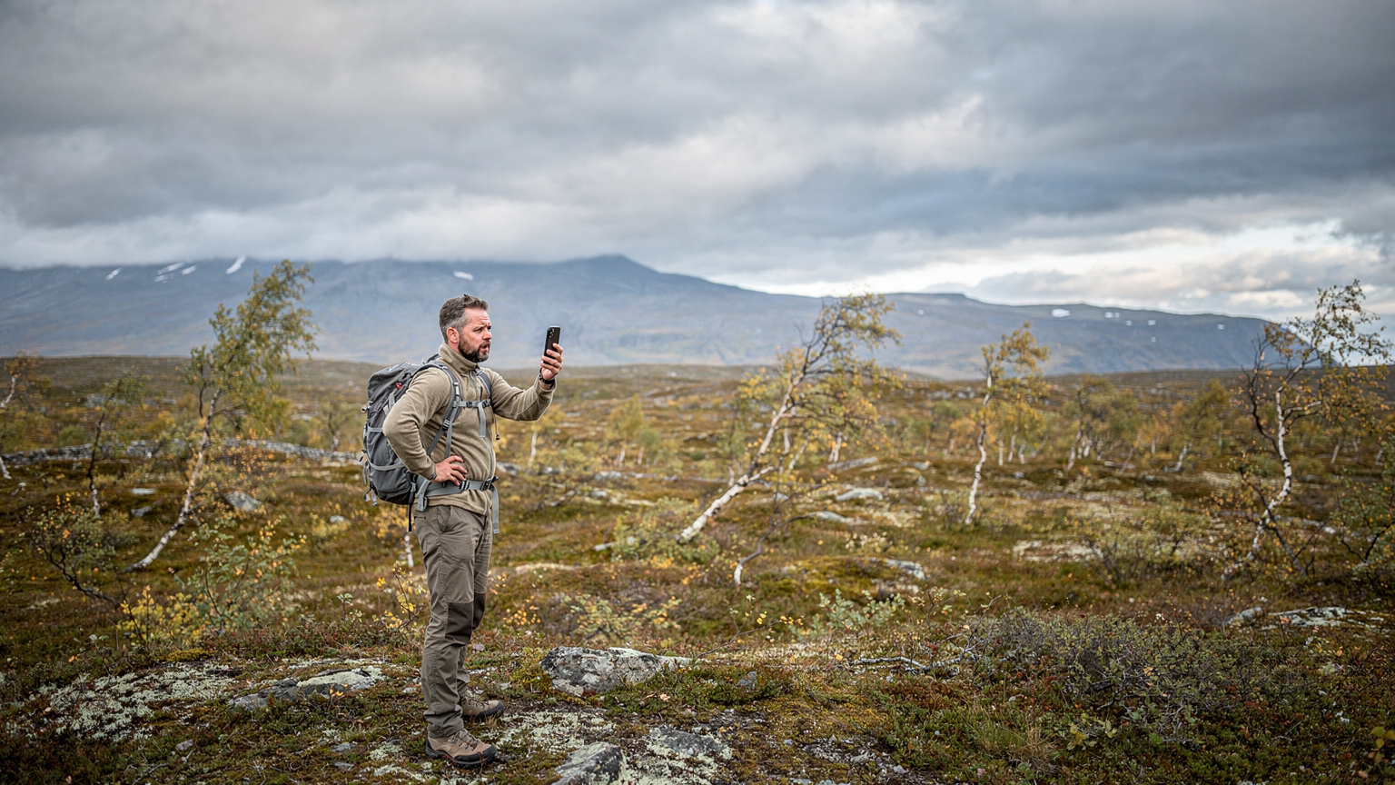 Een gefrustreerde reiziger kijkt naar een smartphone zonder signaal, terwijl hij midden in een uitgestrekt, leeg Scandinavisch landschap staat.