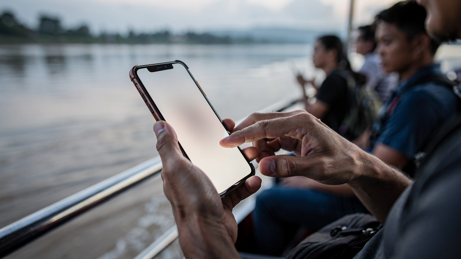 タイとラオスの国境、メコン川を渡るフェリーのデッキで、CellesimのeSIMをスマートフォンに設定する旅行者の手元。周囲には他の旅行者もいる。