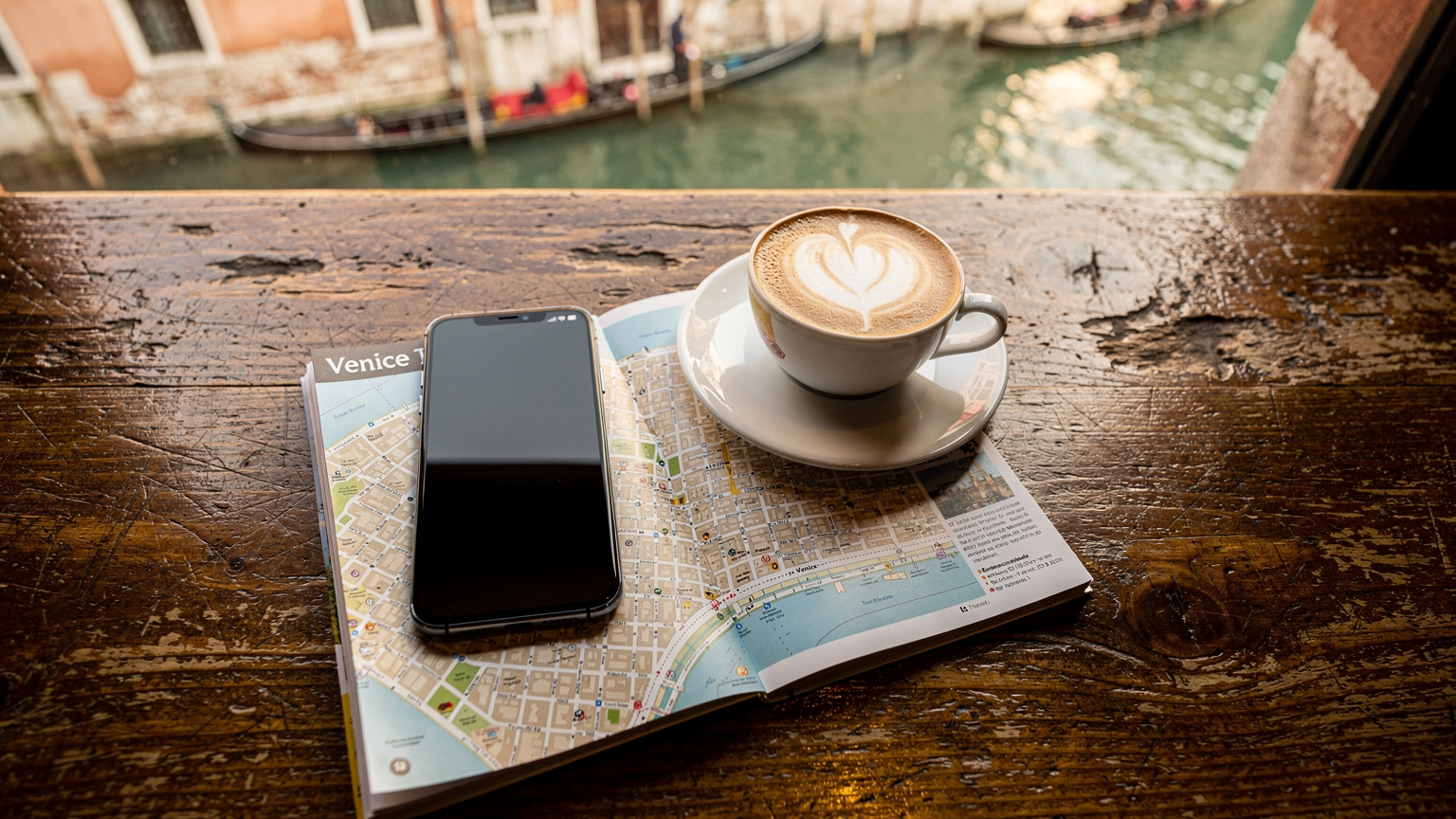 Ein Smartphone liegt auf einer Holztischplatte in einem Café in Venedig, Italien. Neben dem Telefon befinden sich eine kleine Tasse Cappuccino und ein Reiseführer über die Stadt. Im Hintergrund verschwimmen unscharf die geschäftigen Kanäle und Gondeln.