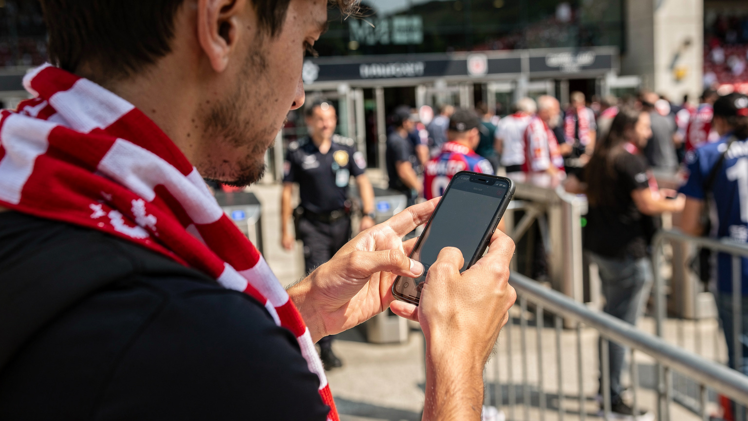 Bir Türk taraftar, Avrupa'da bir stat girişindeki kalabalıkta, telefonunu kullanarak Cellesim uygulamasından maç biletini kontrol ediyor, eSIM sayesinde sorunsuz ve hızlı bağlantı sağlıyor.