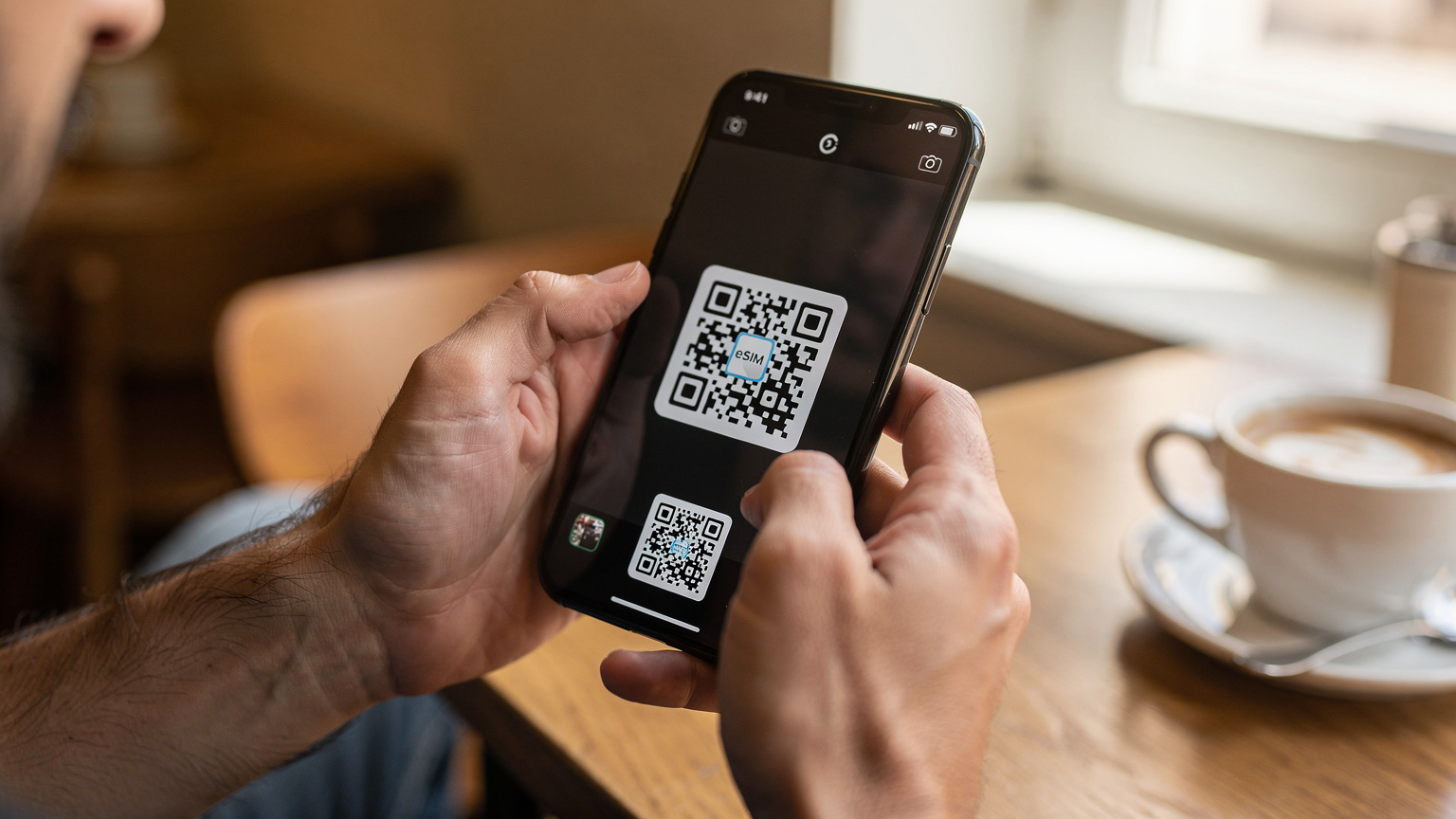 Едър план на ръце, държащи смартфон, докато сканират QR код за активация на eSIM. На заден план се вижда леко разфокусиран интериор на кафене.