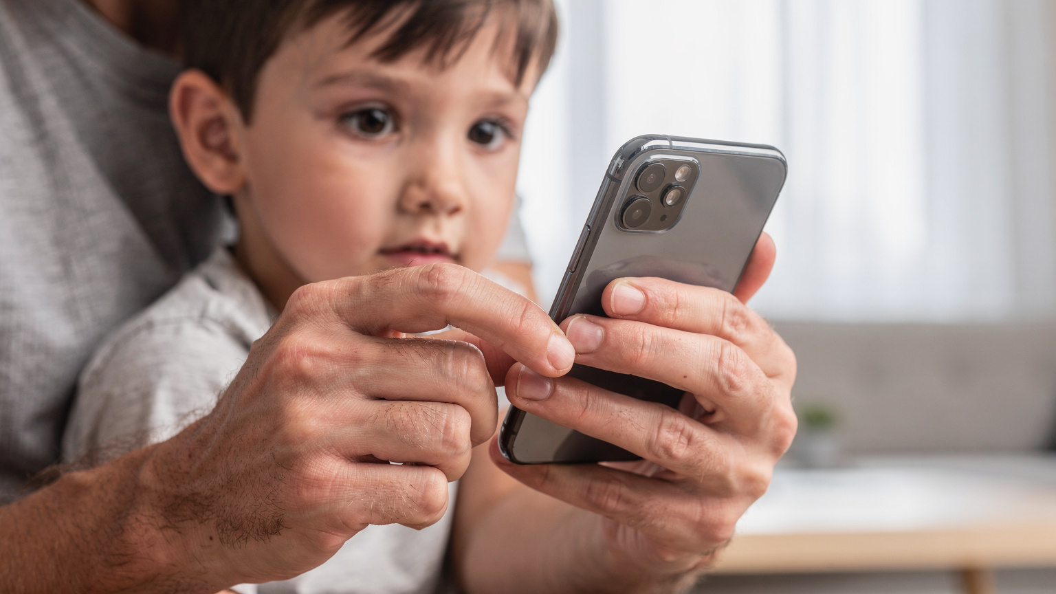 Manos de un padre configurando los ajustes de control parental en un smartphone, con un niño mirando por encima del hombro, en una sala de estar moderna y luminosa.