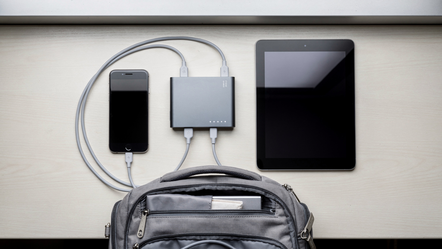 Un power bank de tamaño mediano cargando simultáneamente un smartphone y una tablet, con los cables USB organizados, sobre una superficie limpia de una mochila de viaje abierta.