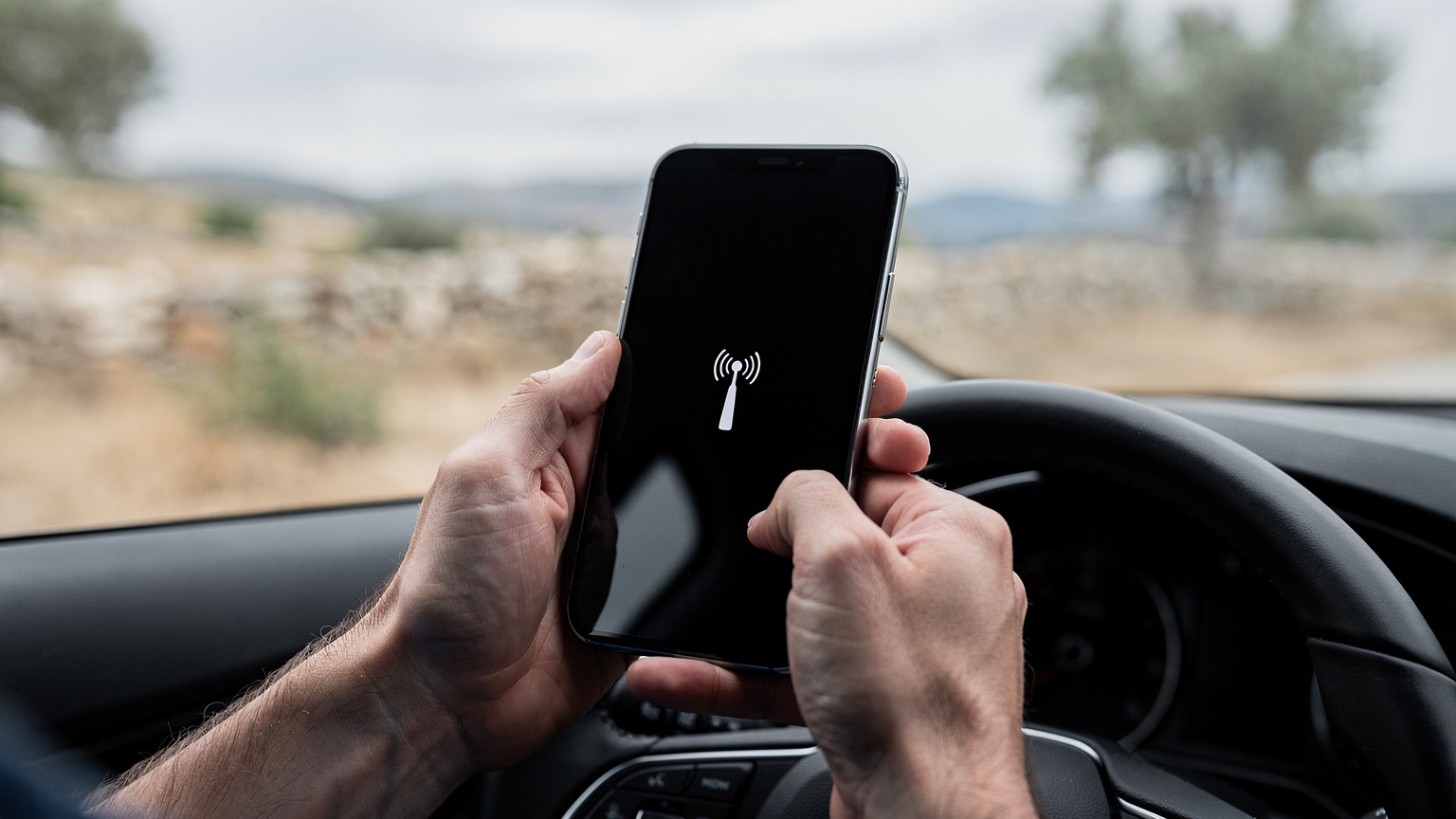 Un smartphone affichant une icône de signal réseau faible, tenu par des mains semblant frustrées dans l'habitacle d'une voiture, avec un paysage rural grec flou en arrière-plan, suggérant une zone isolée sans bonne couverture.