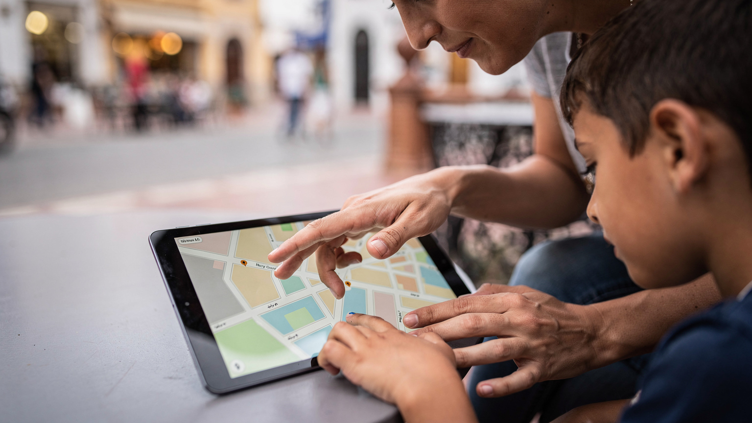 Ręce matki i dziecka trzymające tablet, na którym wyświetla się mapa, z rozmyciem w tle ulicy Tangeru.