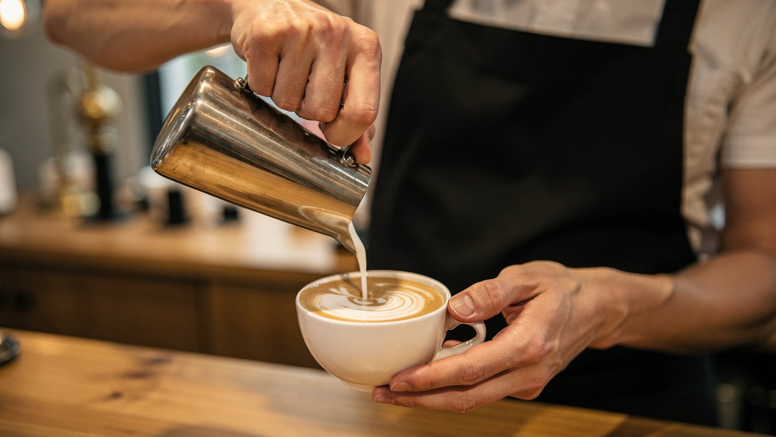 Eine Nahaufnahme zeigt die Hände eines Baristas, der sorgfältig Latte Art in eine Tasse Kaffee gießt, in einem eleganten Café in Südkorea.