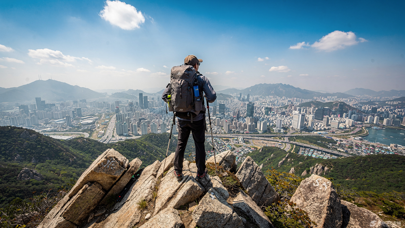 Ein Wanderer genießt die Aussicht von einem Berggipfel im Bukhansan-Nationalpark, mit Seoul im Hintergrund unter einem klaren Himmel.
