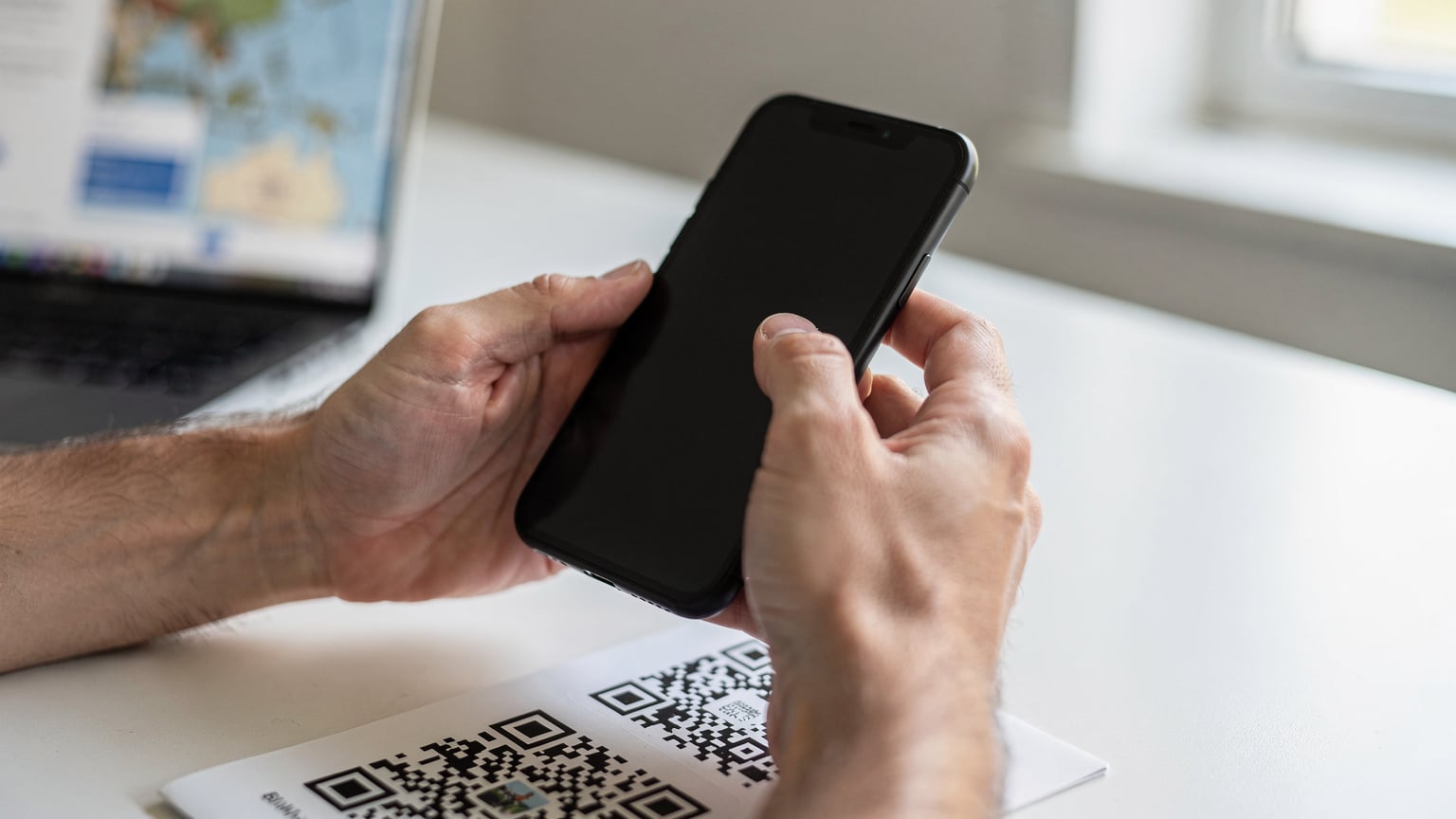 Nahaufnahme der Hände einer Person, die ein Smartphone hält und einen QR-Code scannt, um eine eSIM zu aktivieren. Im Hintergrund ist ein Laptop mit einer geöffneten Reiseplanungs-Webseite zu sehen, vermutlich ein Kartendienst oder eine Unterkunftsseite.