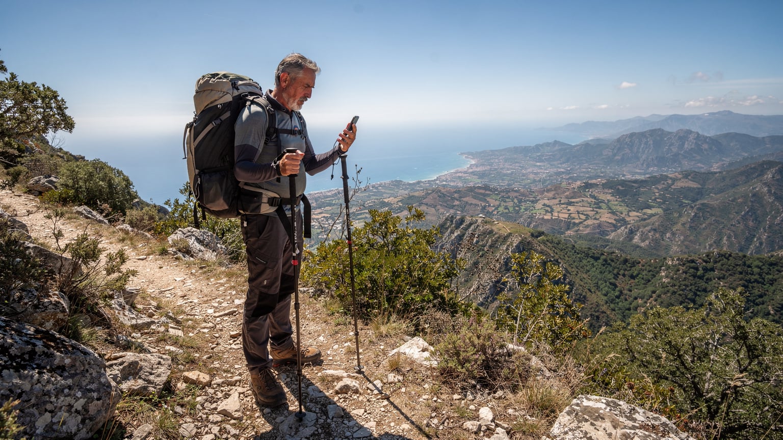 Cestovatel s batohem a turistickými holemi kontroluje signál na smartphonu na odlehlé stezce v horách Sicílie, s panoramatickým výhledem na středomořskou krajinu v pozadí.
