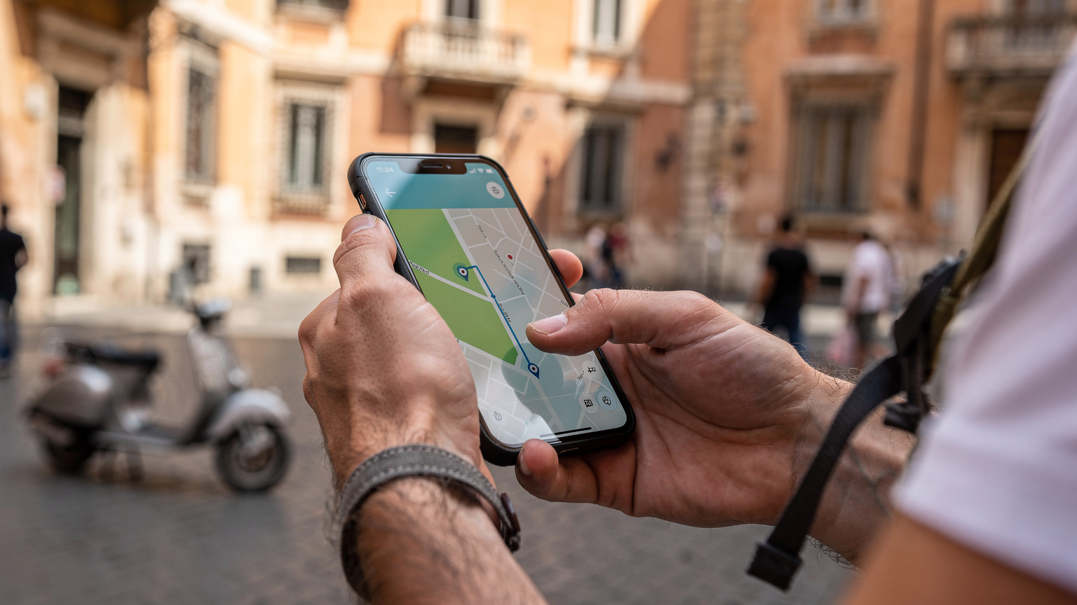 Detailní záběr rukou cestovatele používajícího smartphone k vyhledání trasy na Google Maps, s pozadím rušné římské ulice a historickými budovami.
