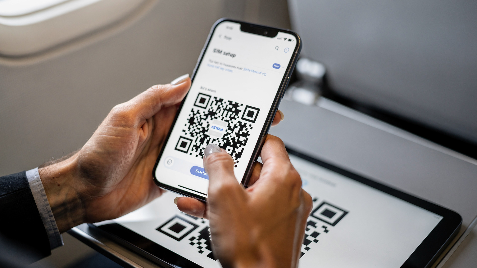 Ръце на човек, държащ смартфон, показващ екрана за настройки на eSIM, докато сканира QR код от друг екран или разпечатка на фона на самолетна седалка.