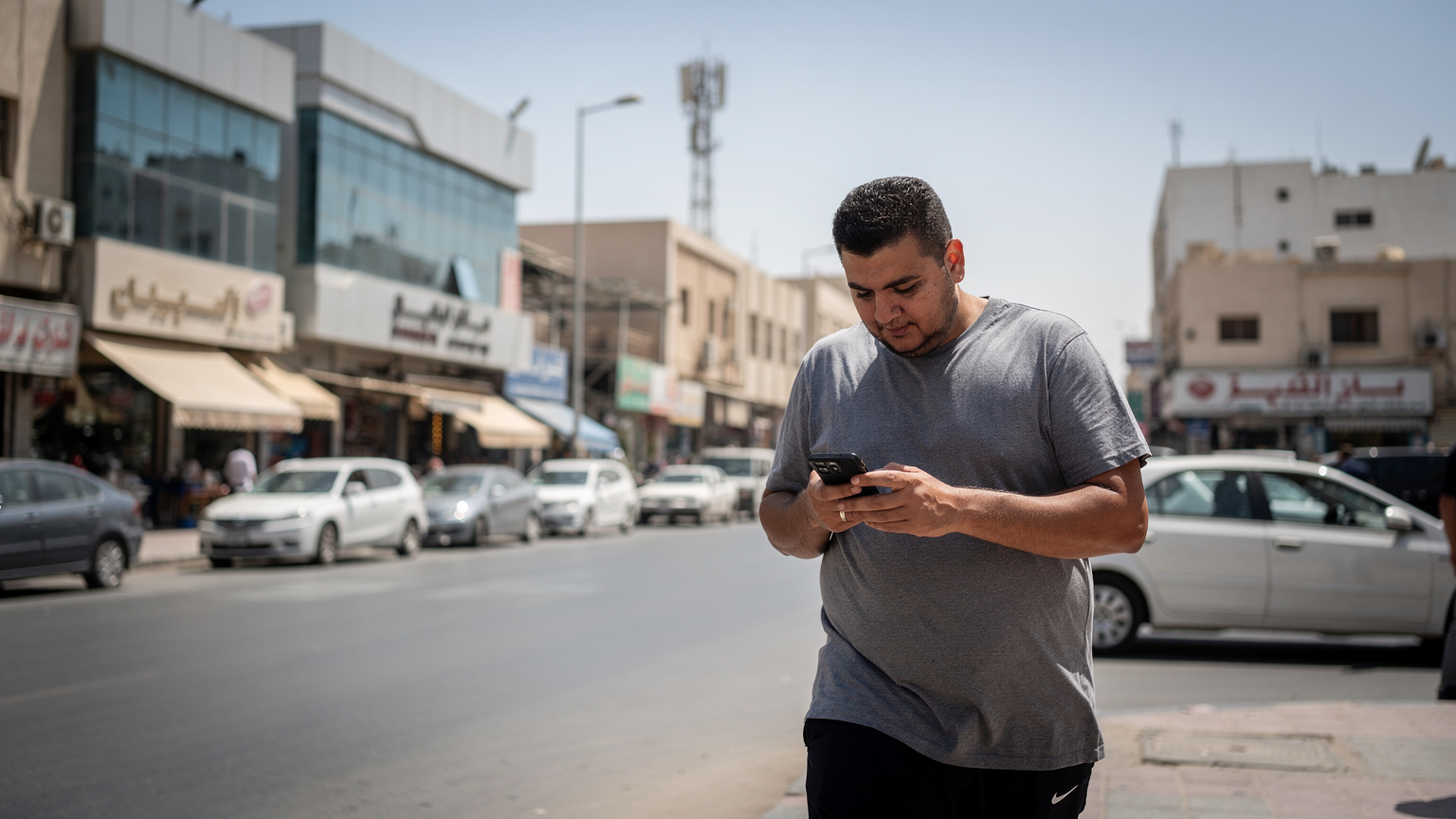 Seorang pejalan kaki menggunakan smartphone-nya di sebuah area perkotaan yang ramai di Jeddah, Arab Saudi, dengan menara telekomunikasi modern terlihat samar di kejauhan.