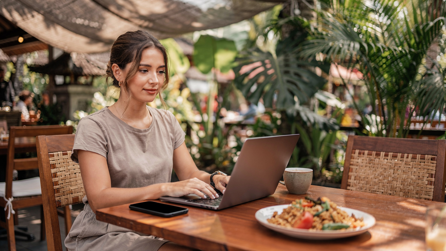 A digital nomad works on a laptop at a cafe in Ubud, Bali, with a plate of local food and a phone nearby, indicating a balance of work and local experience.