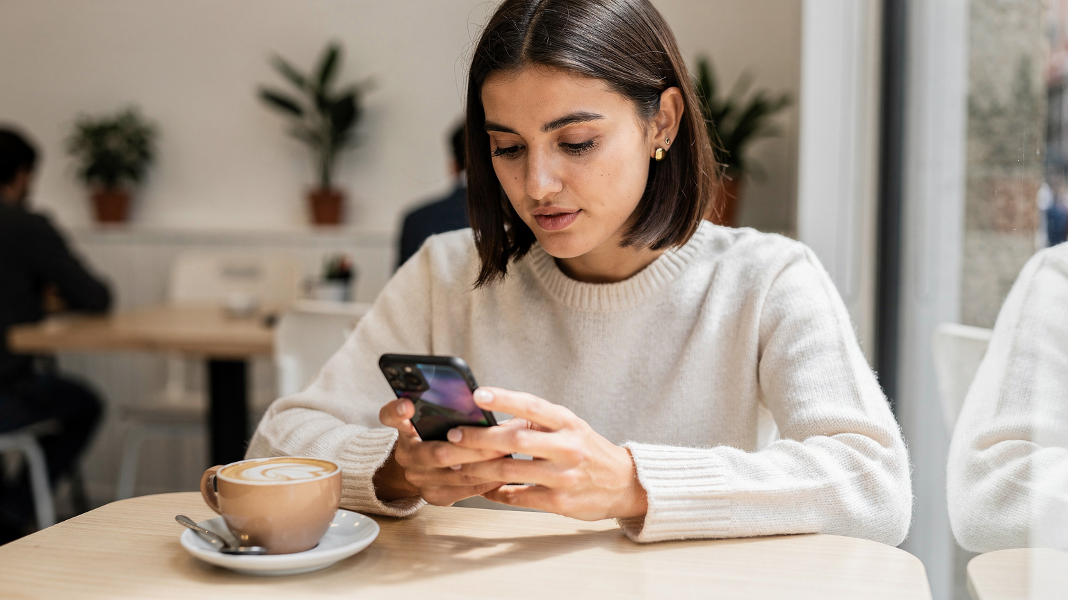 Una mujer española joven, usando su smartphone, activa una eSIM mientras está sentada en un moderno café de Madrid, con un café con leche sobre la mesa.