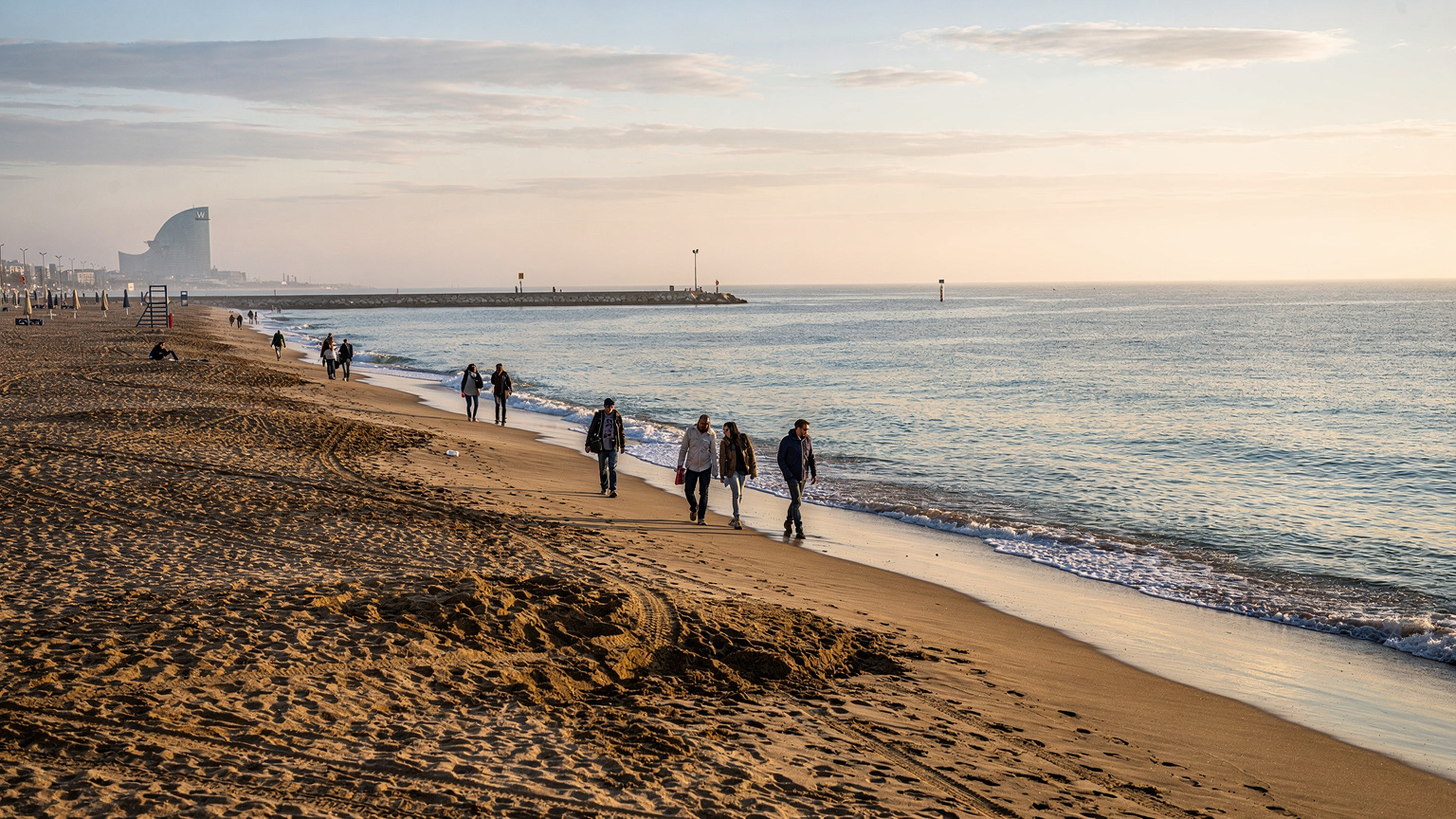Una veduta panoramica della spiaggia della Barceloneta al mattino presto, con poche persone che passeggiano e il mare calmo, suggerendo una tranquillità prima dell'affollamento turistico.