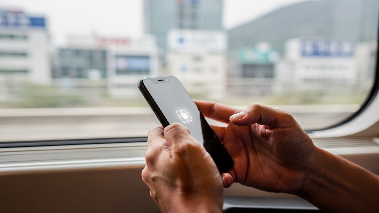 Manos de un viajero utilizando un smartphone con una eSIM activa a bordo de un tren KTX de alta velocidad, con el paisaje urbano de Corea del Sur pasando por la ventana.