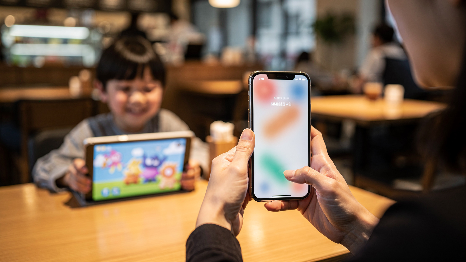 韓国ソウルのカフェで、母親がiPhoneのeSIM設定画面でデータ使用量を確認し、子どもがタブレットで動画を見ている様子。