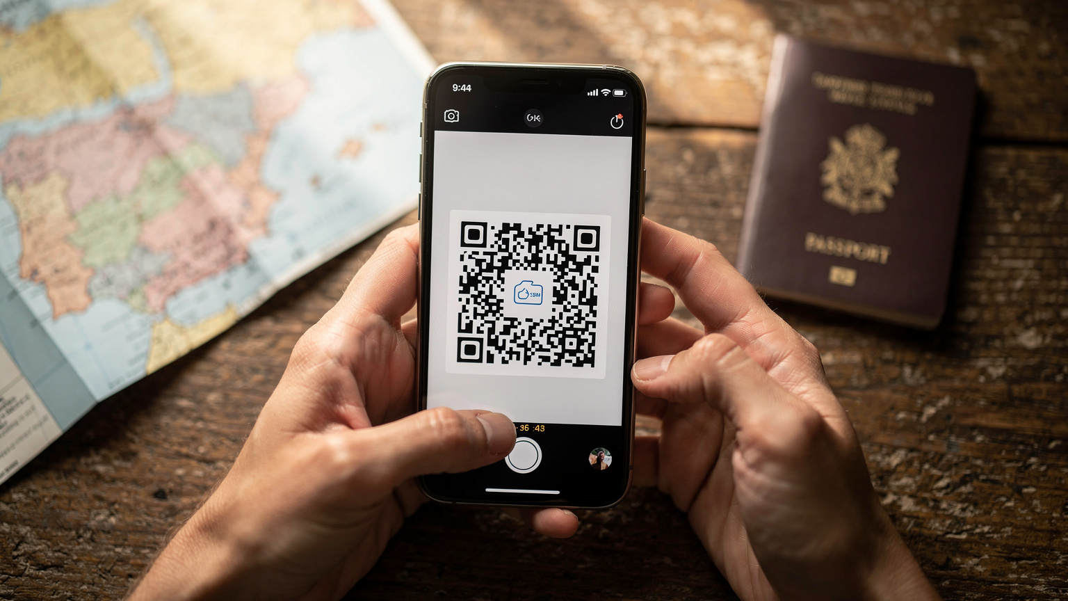 Manos de una persona sosteniendo un smartphone, mostrando el proceso de escaneo de un código QR para activar una eSIM. El fondo está suavemente desenfocado, con motivos de viaje como un mapa y un pasaporte sobre una mesa de madera.