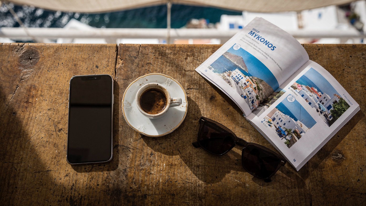 Una mesa de café en Little Venice, Mykonos, con un smartphone, una taza de café griego, unas gafas de sol y una guía de viaje, listos para un día de exploración.