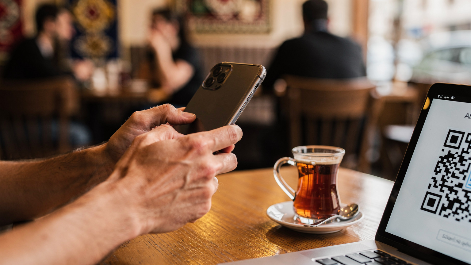 Bir kafede oturan kişinin akıllı telefonunda bir QR kodunu tararken görünen elleri. Arka planda Ankara'nın hareketli bir caddesi ve bir kaç cafe dışarı masası.