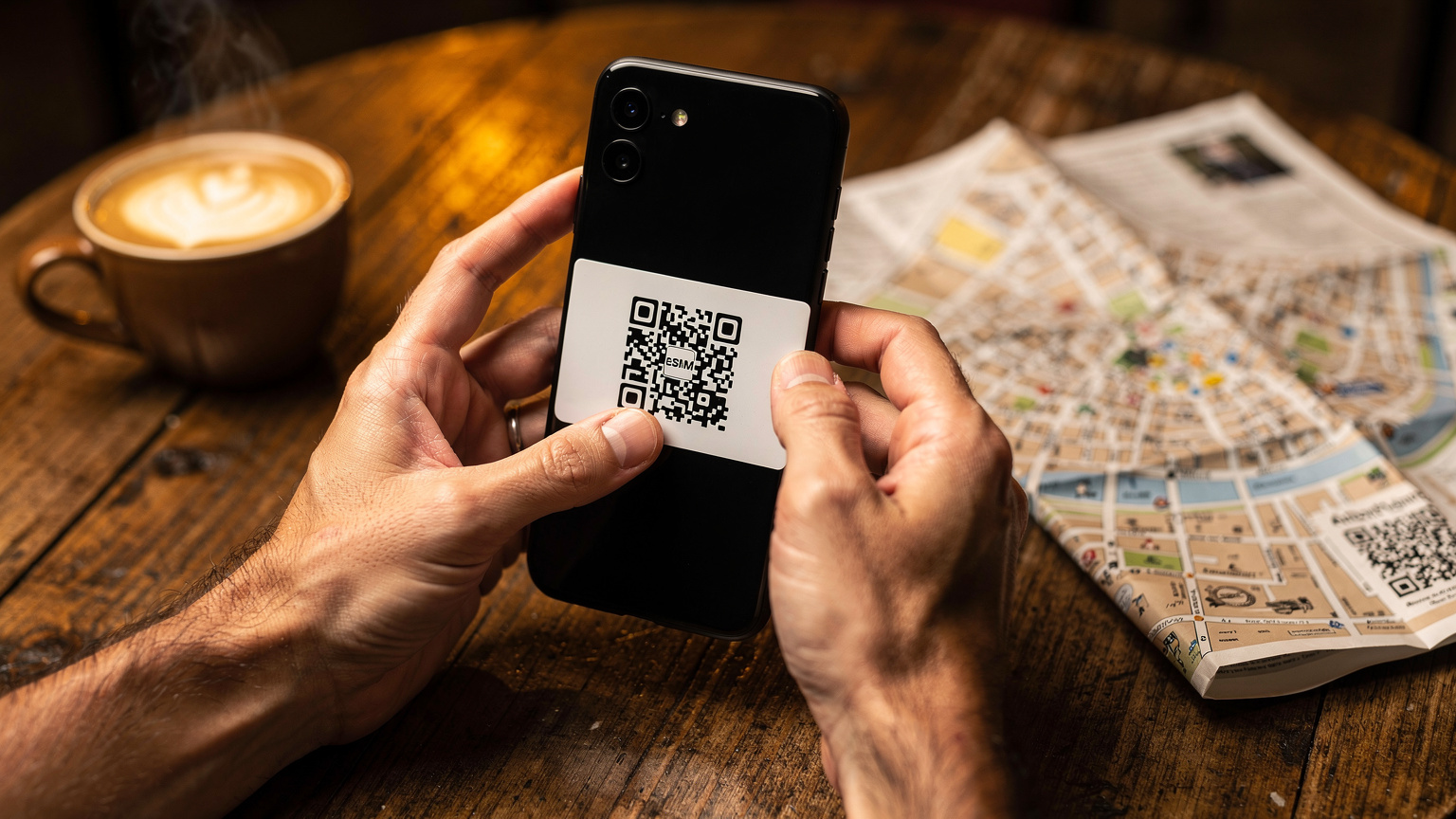 Primer plano de unas manos sosteniendo un smartphone mientras se escanea un código QR para activar una eSIM en una mesa de madera de un café, con una taza de café humeante y un mapa de papel borroso de fondo. El ambiente es acogedor y rústico.