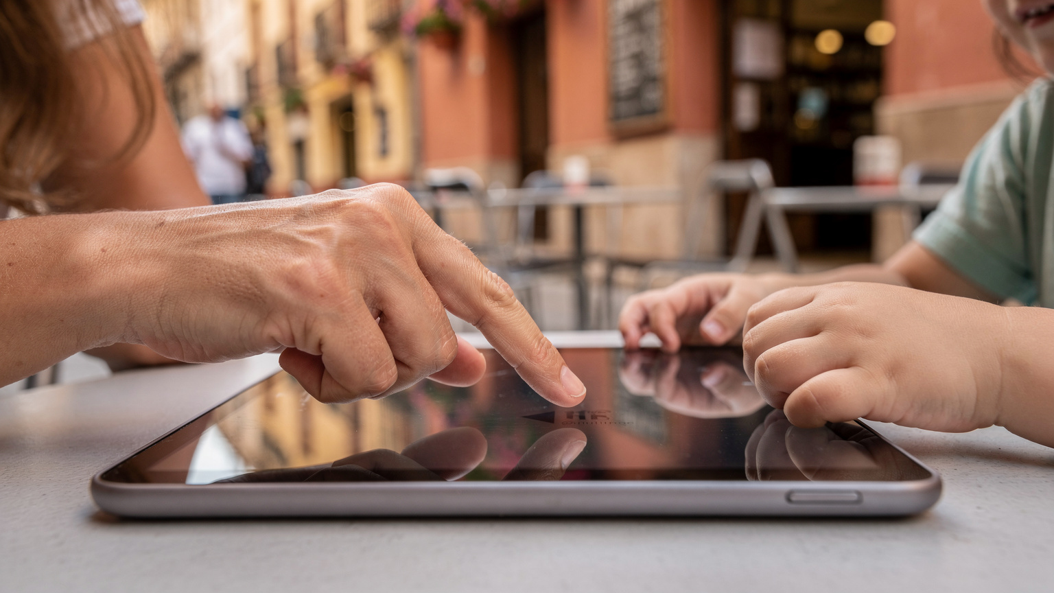 Bir annenin eli, çocuğunun elindeki tabletin ekranında ebeveyn kontrol ayarlarını yaparken görünüyor, arka planda bulanık bir İspanyol sokağı.