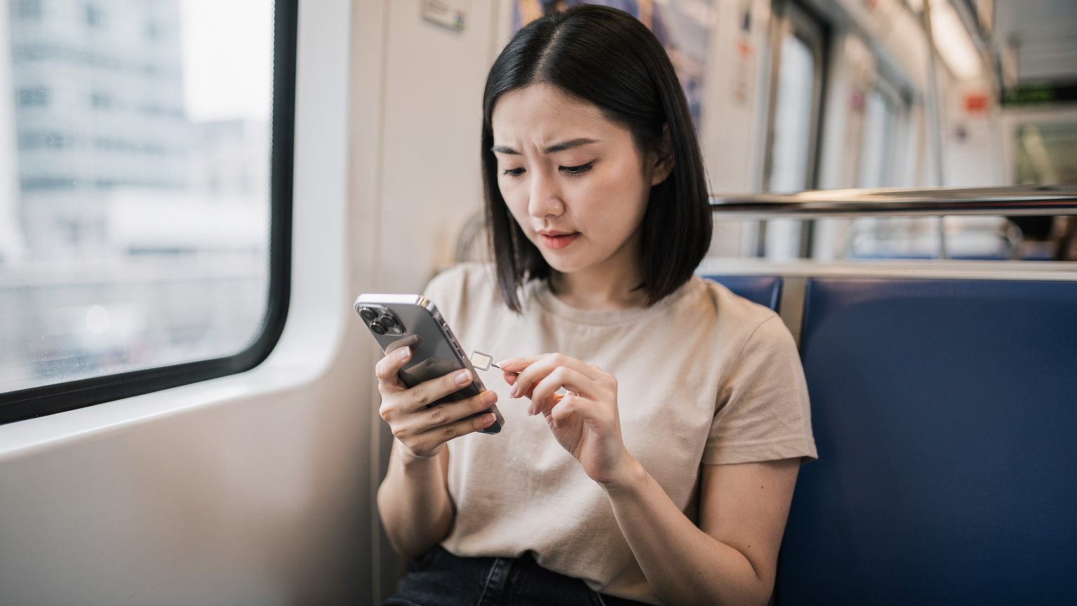 一位香港年輕女性在地鐵車廂內檢查iPhone的SIM卡槽,專注地查看是單實體卡還是雙實體卡版本。