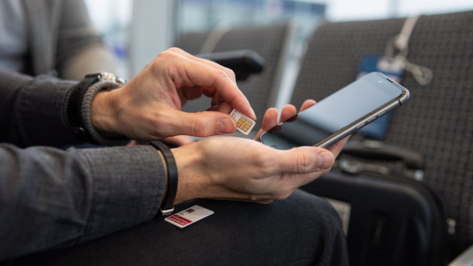 Rukou cestovatele drží starou fyzickou SIM kartu, zatímco se snaží vložit novou lokální SIM kartu do slotu telefonu, v rušném prostředí letiště.