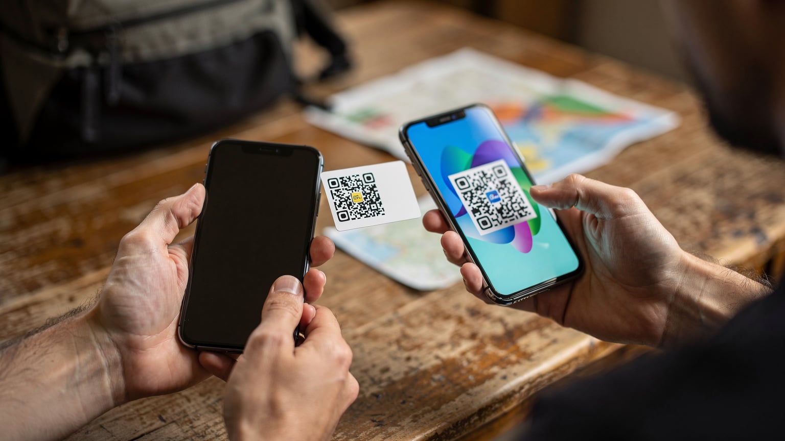 Detailní záběr rukou cestovatele s batohem, jak drží chytrý telefon a skenuje QR kód pro aktivaci eSIM, s rozmazaným batohem a mapou v pozadí.