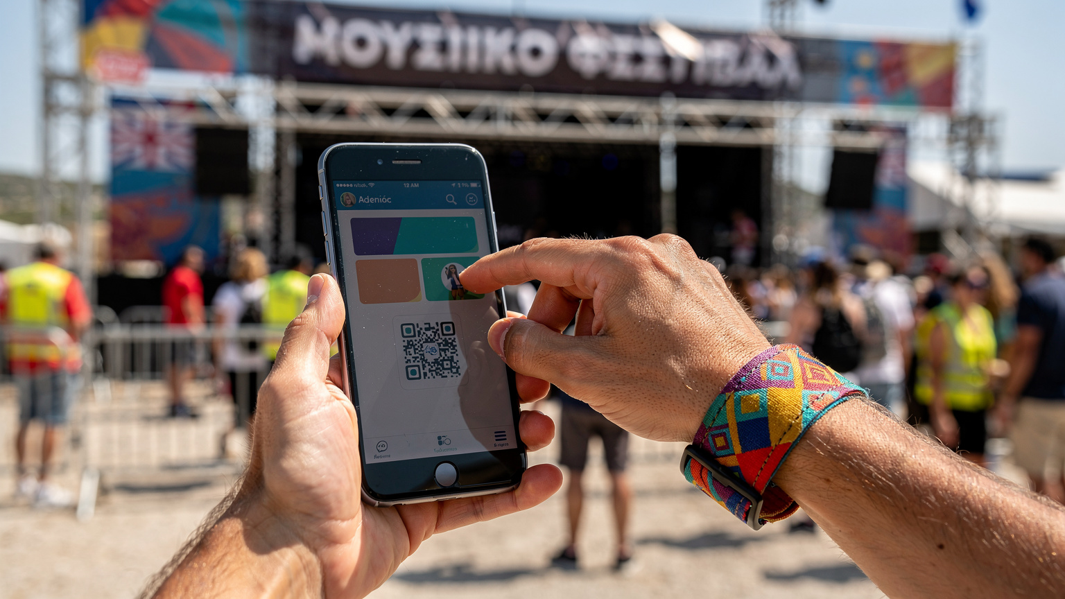 Ένα χέρι κρατά ένα smartphone που δείχνει μια εφαρμογή εισιτηρίων φεστιβάλ, με ένα άλλο χέρι να χτυπάει ένα βραχιόλι εισόδου στο φεστιβάλ, με φόντο έναν πολυσύχναστο είσοδο φεστιβάλ στην Ελλάδα.