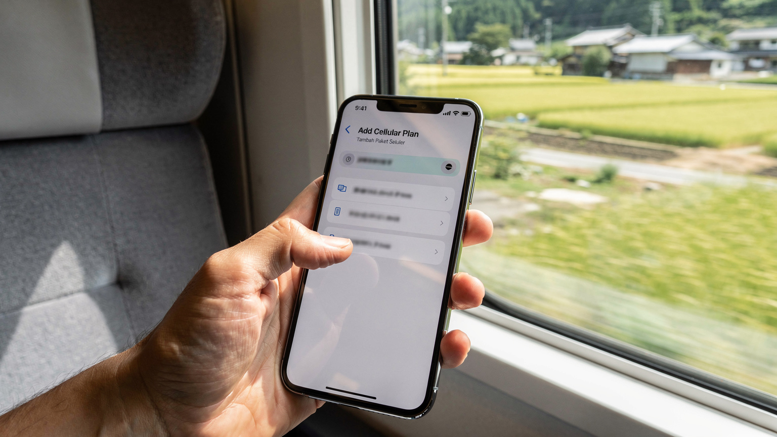 Sebuah tangan memegang smartphone menampilkan layar pengaturan seluler dengan opsi 'Tambah Paket Seluler' yang sedang dipilih, sementara di latar belakang terlihat kereta Shinkansen melaju cepat di pedesaan Jepang.