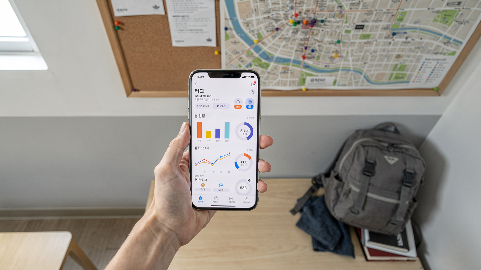 Sebuah tangan manusia memegang smartphone yang menampilkan aplikasi pemantau penggunaan data seluler di layar, di dalam sebuah kamar hostel yang rapi di Seoul, Korea Selatan, dengan peta kota di dinding dan ransel di sudut.