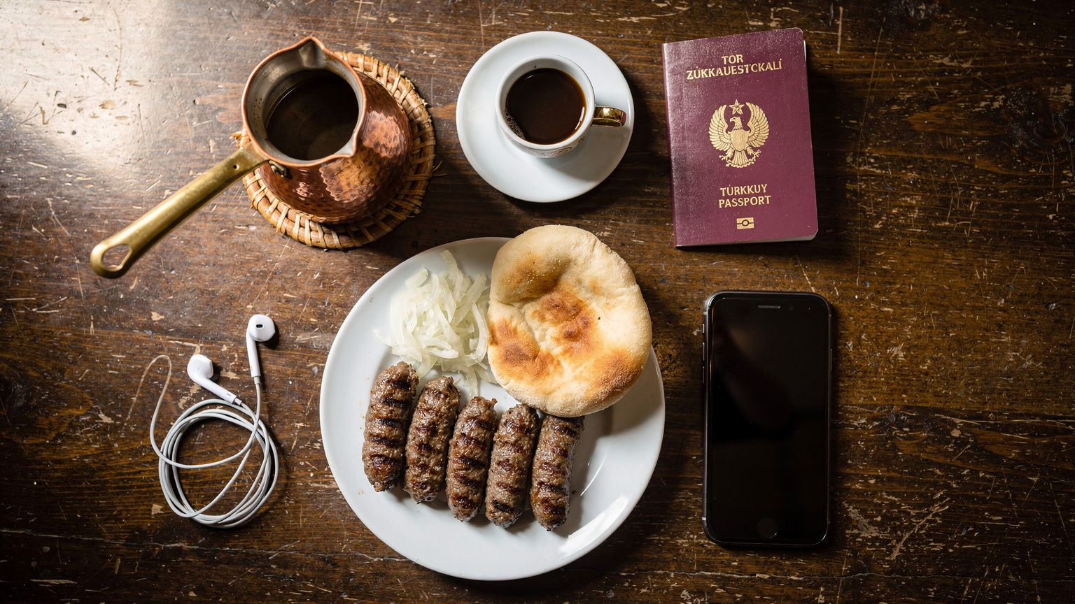 Bir Saraybosna kafesinde, bir masanın üzerinde yerel kahve fincanı, bir adet Cevapi tabağı, pasaport, telefon ve kulaklıkların olduğu bir overhead flat-lay fotoğrafı.