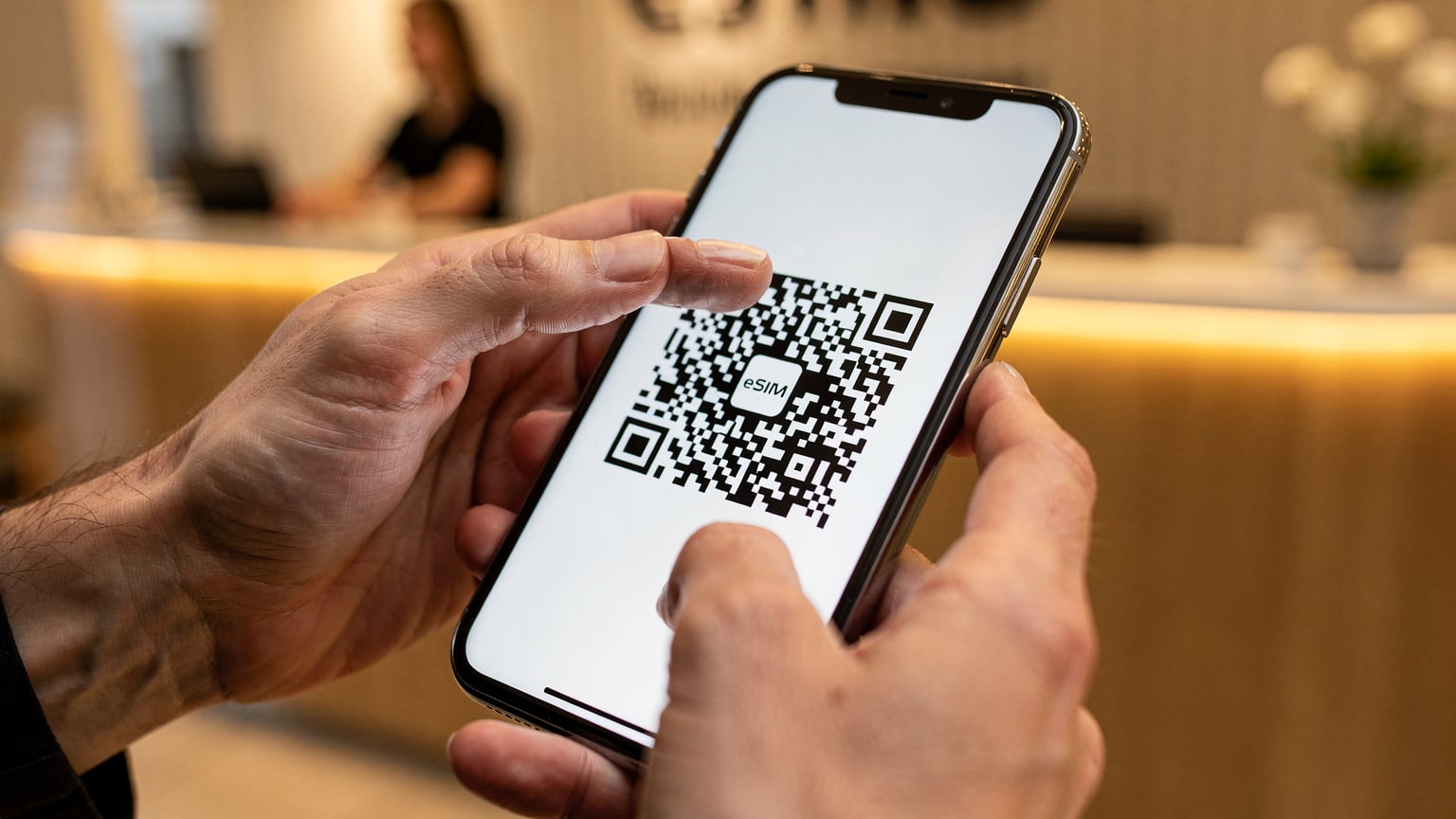 Nahaufnahme von Händen, die ein Smartphone halten, auf dessen Bildschirm ein QR-Code für die eSIM-Aktivierung angezeigt wird, im Hintergrund eine leicht verschwommene Hotelrezeption.