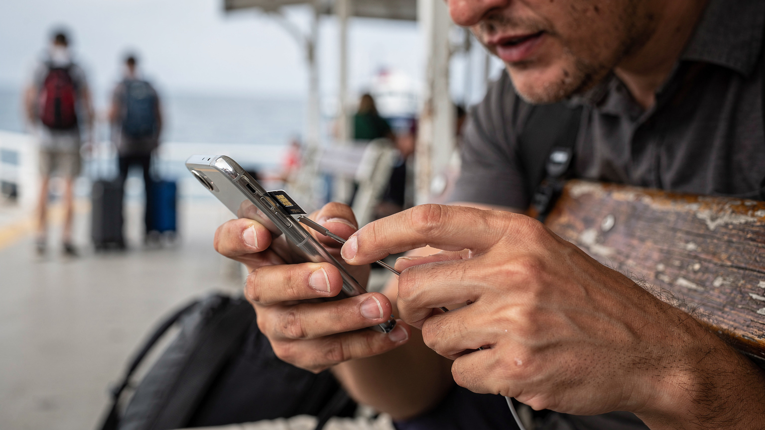 Nærbillede af en rejsende, der forsøger at indsætte et lille, fysisk SIM-kort i en telefon, mens de sidder på en træbænk ved en færgeterminal, med et frustreret udtryk i ansigtet.