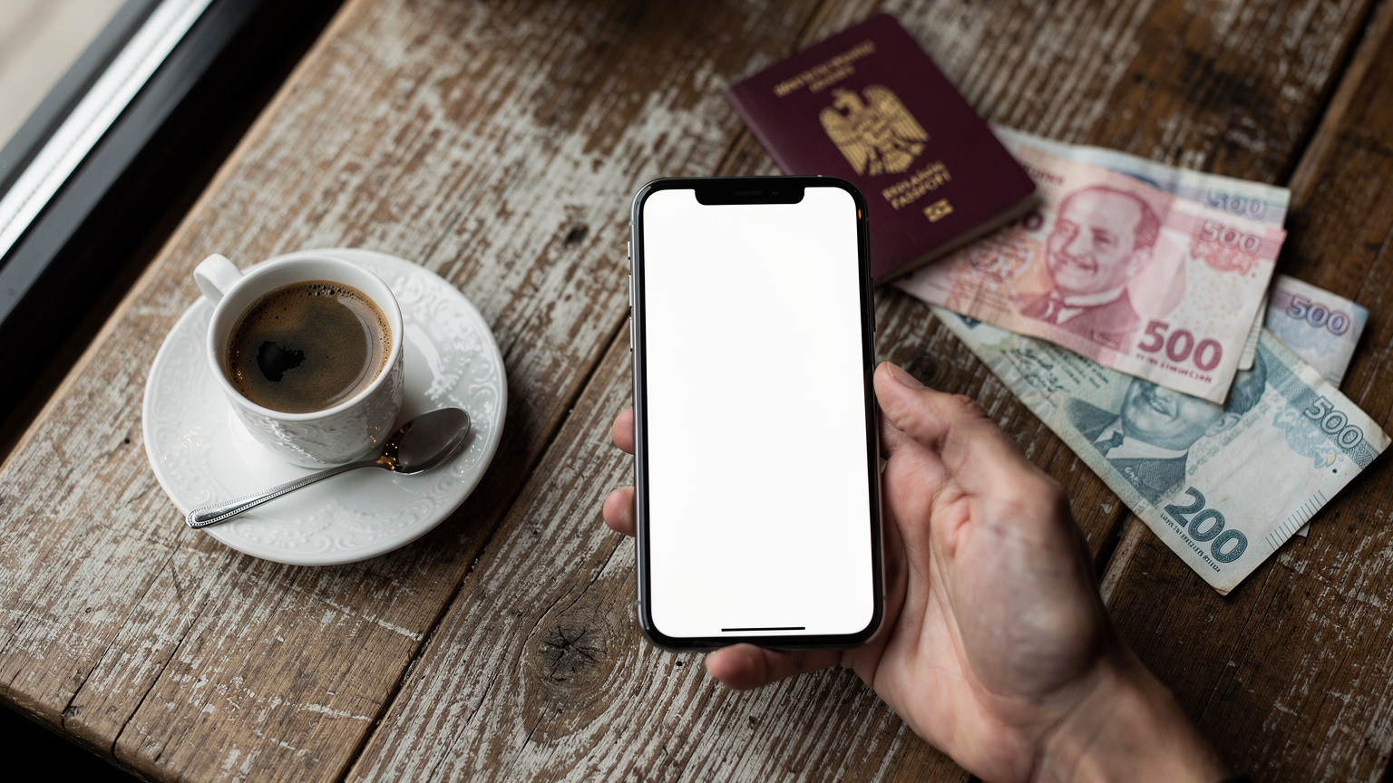 O persoană ține un smartphone în mână, arătând un ecran curat, fără text vizibil, în timp ce pe masă, lângă o ceașcă de cafea turcească, se află un pașaport românesc și câteva lire turcești. Scena este într-o cafenea modernă din Istanbul.