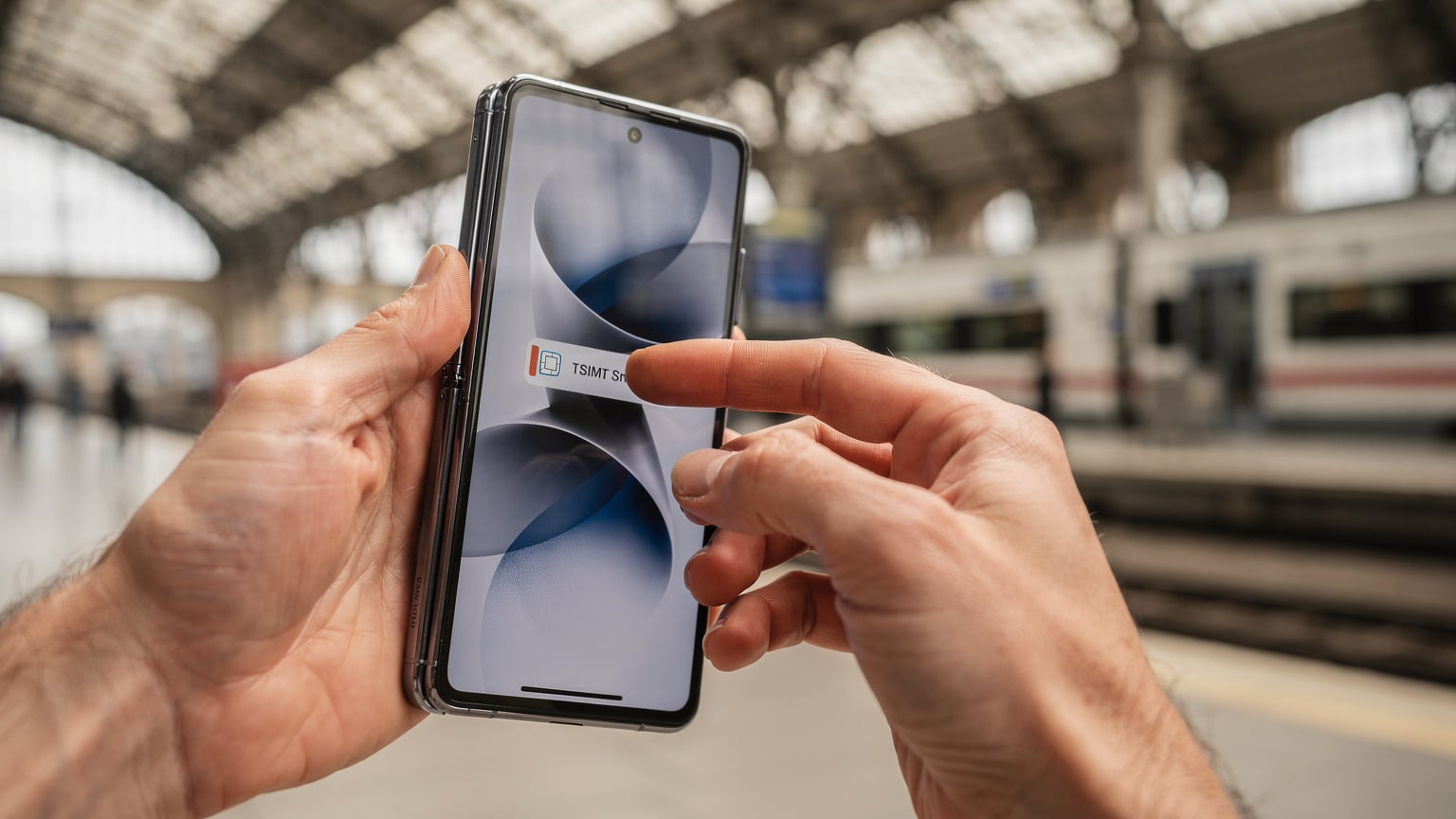 Detailní záběr na ruce, které plynule přepínají mezi eSIM profily na Samsungu Galaxy Z Fold6, s rozmazaným pozadím typického evropského nádraží.