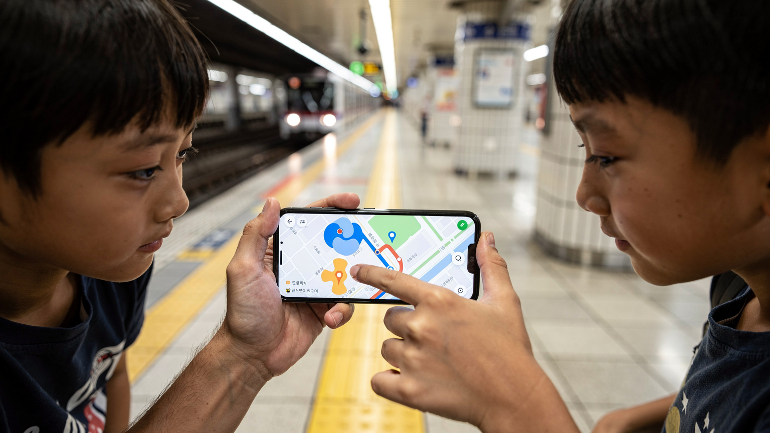 Tangan seorang bapa menunjukkan destinasi di peta digital pada telefonnya kepada dua orang anak yang melihat dengan penuh minat, di sebuah stesen kereta api bawah tanah yang sibuk di Seoul, Korea Selatan.