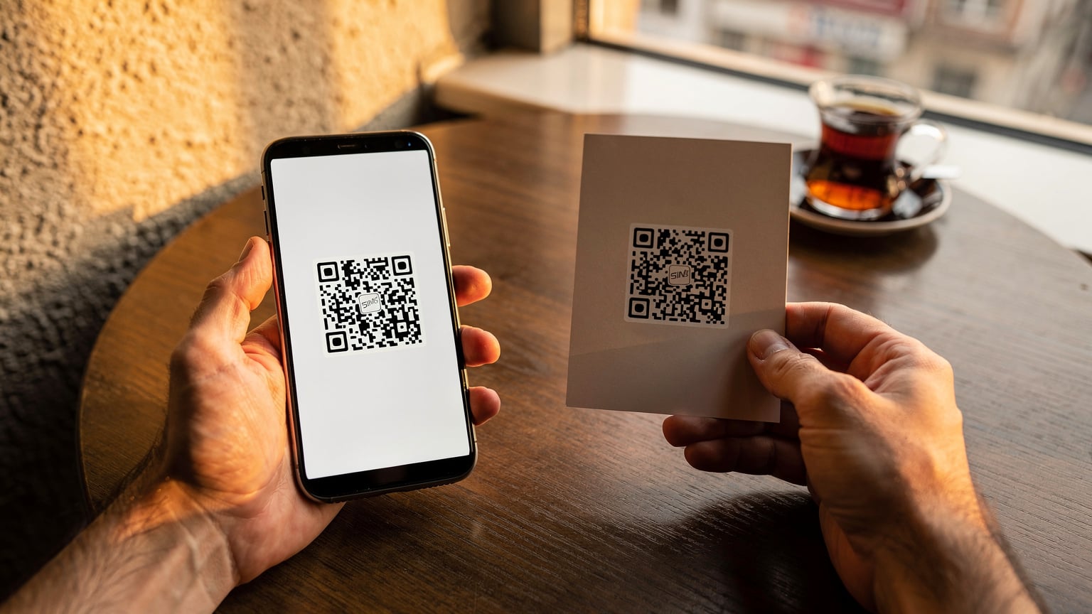 O mână ține un telefon mobil pe care se afișează un cod QR, iar cealaltă mână scanează codul de pe o broșură, în timp ce lumina caldă a soarelui intră printr-o fereastră, sugerând o activare ușoară și rapidă a unui eSIM.