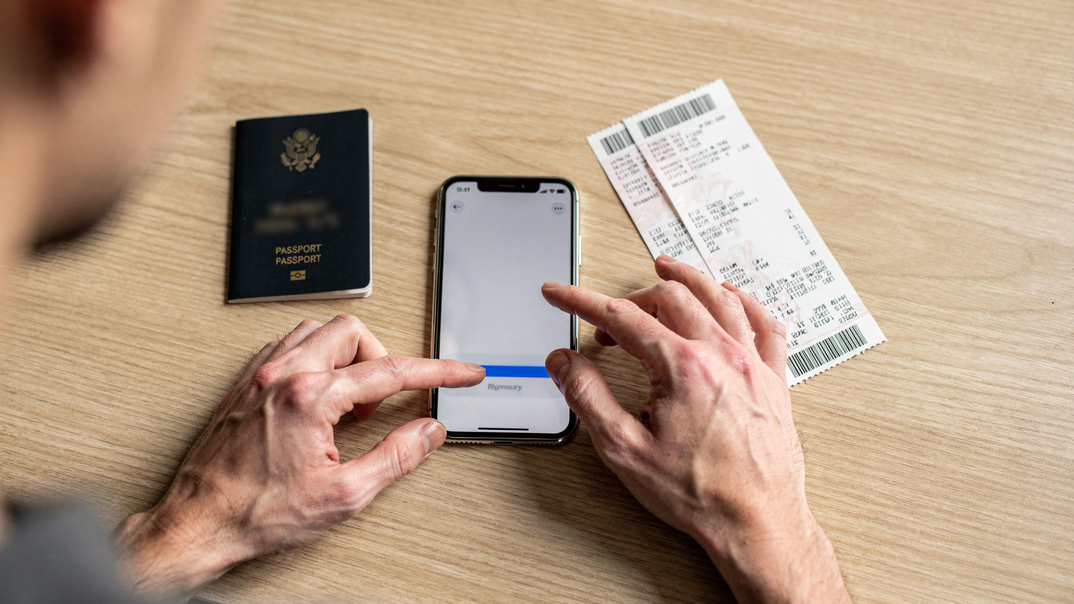 旅行前の自宅で、パスポートや航空券、iPhoneをテーブルに並べ、eSIMの購入と設定を準備している旅行者の手元。指先でiPhoneの画面を操作している。